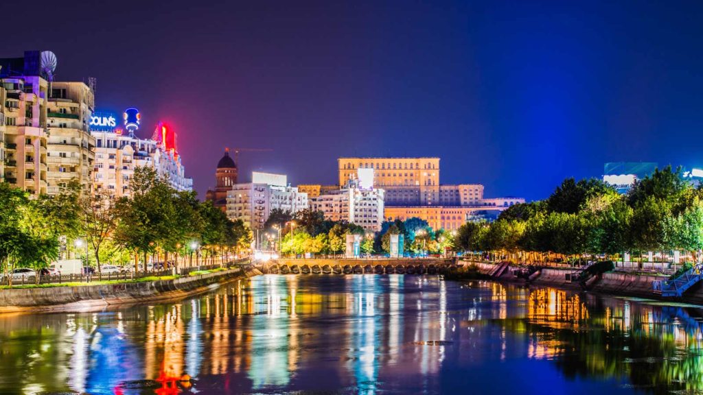 Nachtaufnahme einer Stadt mit beleuchteten Gebäuden und einem Fluss im Vordergrund, der die Lichter reflektiert.