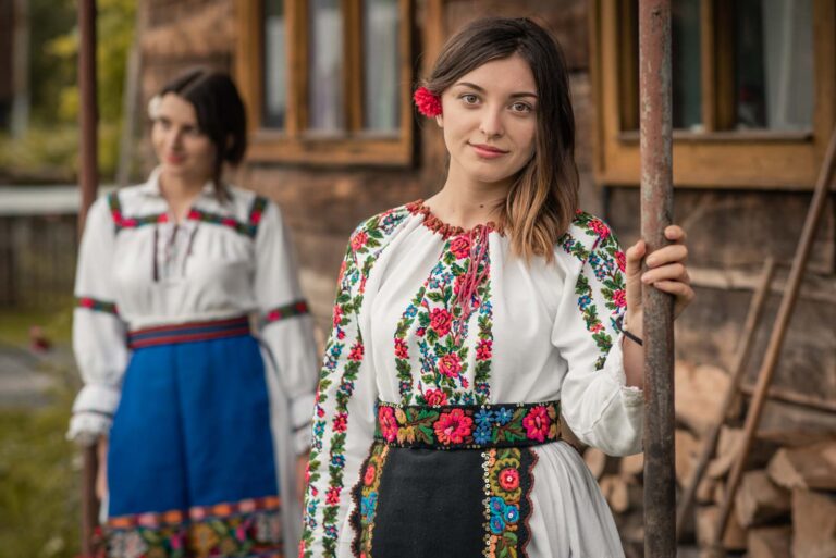 Zwei Frauen tragen traditionelle Kleidung mit bunten Stickmustern; die eine Frau steht im Vordergrund und hält eine Stange, während die andere im Hintergrund verschwommen zu sehen ist.