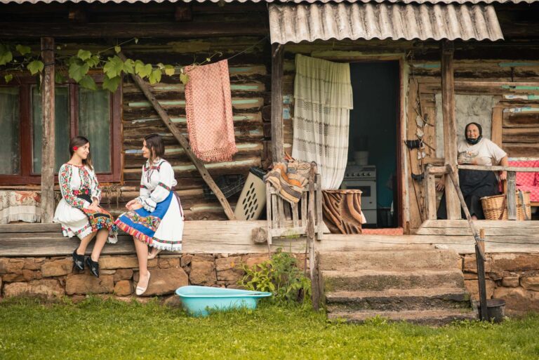 Zwei Frauen in traditioneller Kleidung sitzen auf der Veranda eines alten Holzhauses, während eine ältere Frau im Hintergrund auf einem Stuhl sitzt.