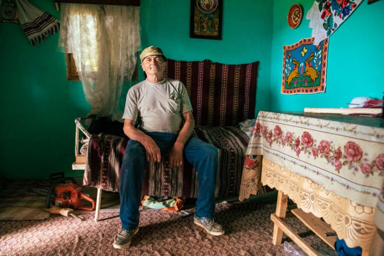 Ein älterer Mann sitzt in einem traditionell eingerichteten Raum mit grünen Wänden, verziert mit farbenfrohen Stickereien. Er trägt ein helles T-Shirt, eine blaue Hose und eine Mütze. Neben ihm steht eine Kettensäge auf dem Boden.