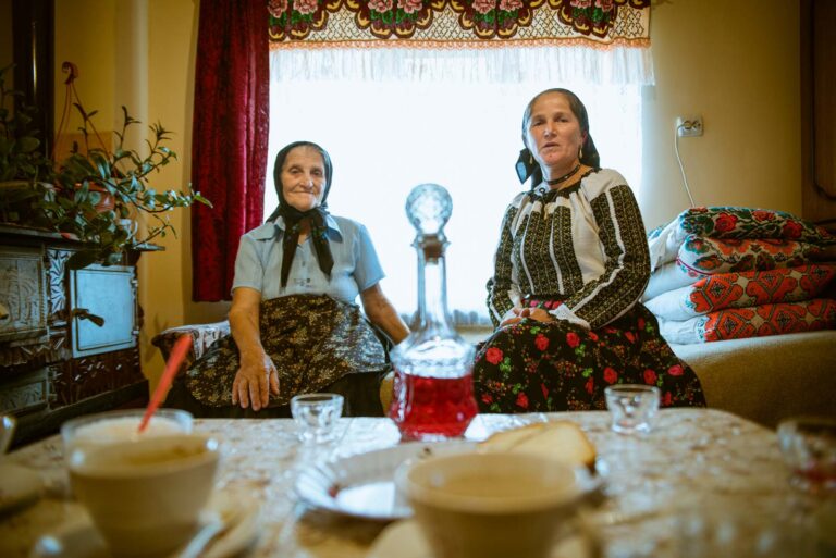 Zwei ältere Frauen in traditioneller Kleidung sitzen in einem Wohnzimmer auf einer Bank; im Vordergrund ein Tisch mit Getränken und Geschirr, im Hintergrund ein Fenster mit roten Vorhängen.