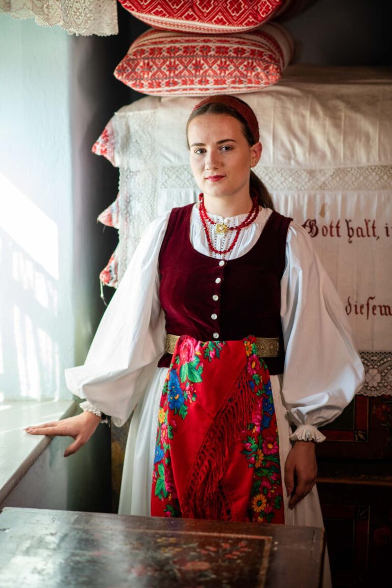 Frau in traditioneller Tracht mit blauer Weste, weißer Bluse und rotem, geblümtem Hüfttuch, steht neben einem Tisch vor gestapelten, bestickten Kissen.