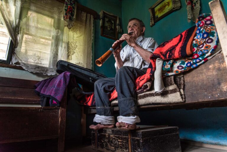 Ein älterer Mann sitzt auf einem Bett in einem Zimmer mit blauen Wänden und spielt eine hölzerne Flöte. Vor ihm liegt ein bunt gemustertes Tuch. Ein Fenster mit Spitzengardinen und ein paar Bilder sind im Hintergrund zu sehen.