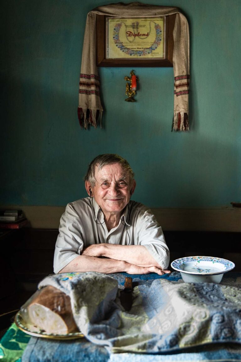 Ein älterer Mann sitzt lächelnd an einem Tisch mit einem bedeckten Brotlaib und einer Schüssel. Hinter ihm hängt ein gerahmtes Diplom an der Wand, dekoriert mit einem Tuch und einer kleinen Kerze in der Halterung darunter.
