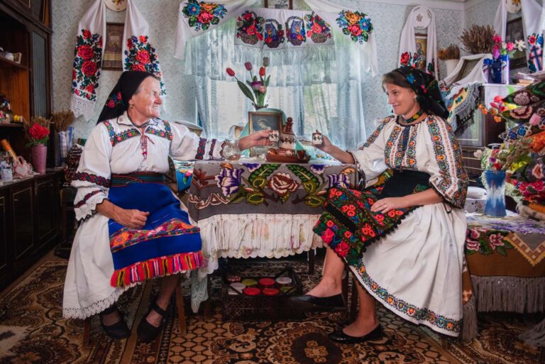 Zwei Frauen in traditioneller Kleidung sitzen in einem Zimmer mit bunten Stickereien und trinken aus kleinen Tassen.