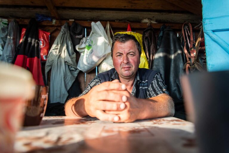Ein Mann sitzt an einem Tisch in einem rustikalen Raum mit verschiedenen aufgehängten Jacken und Taschen im Hintergrund.