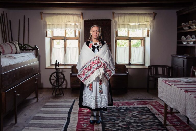 Eine Frau in traditioneller, bestickter Kleidung steht in einem rustikalen, gemütlich eingerichteten Raum mit Holzmöbeln.