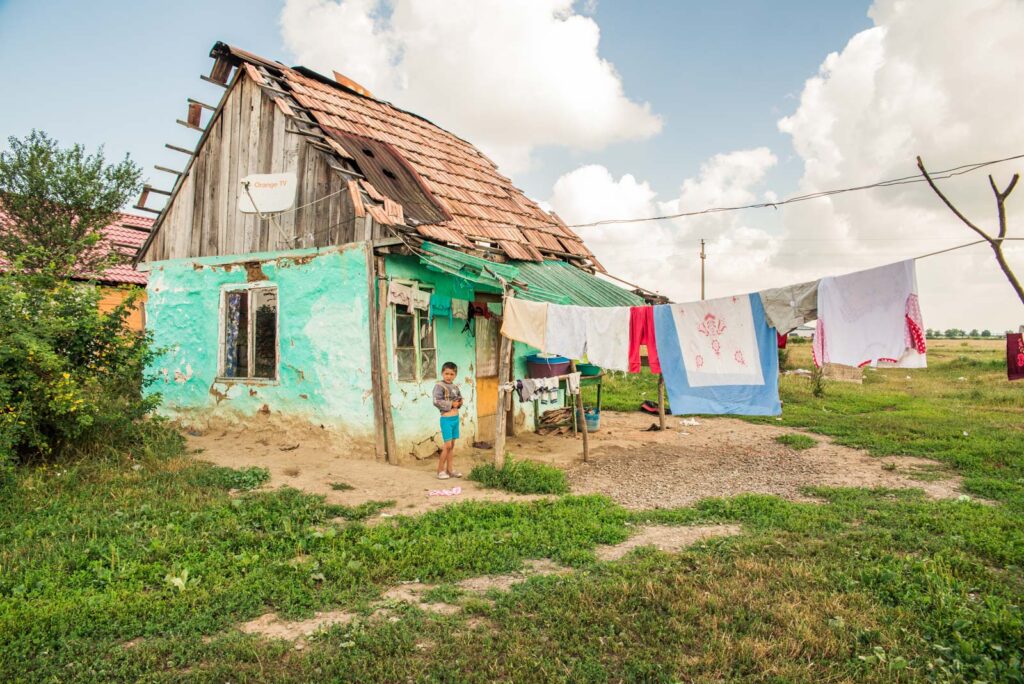 Ein kleines, verfallenes Haus mit beschädigtem Dach und abblätternder Farbe, vor dem ein Kind steht. Neben dem Haus hängt Wäsche auf einer Leine. Der Himmel ist bewölkt, und das Haus steht auf einer grasbewachsenen Fläche.