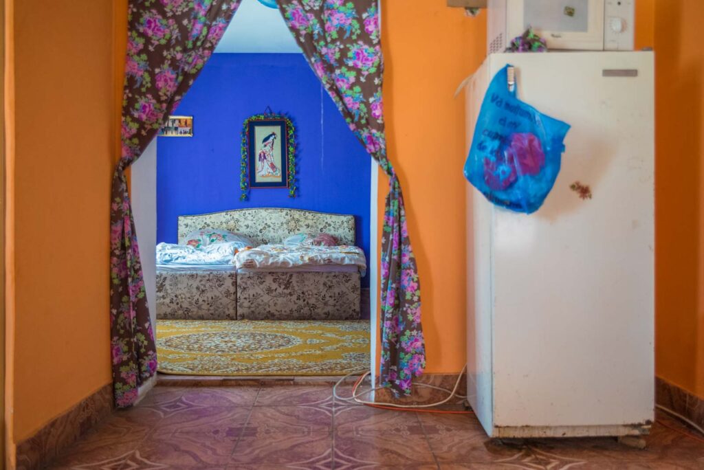 Innenraum mit bunter Einrichtung; im Vordergrund ein Kühlschrank neben einem Vorhang mit Blumenmuster, dahinter ein blau gestrichener Raum mit einem Bett und einem an der Wand hängenden Bild.
