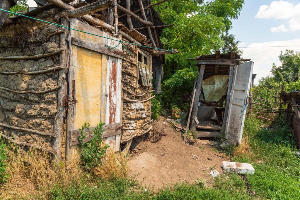 Eine verlassene, hölzerne Hütte mit zerfallenen Wänden und Dach, daneben eine alte, schiefe Toilette, umgeben von Gras und Bäumen.