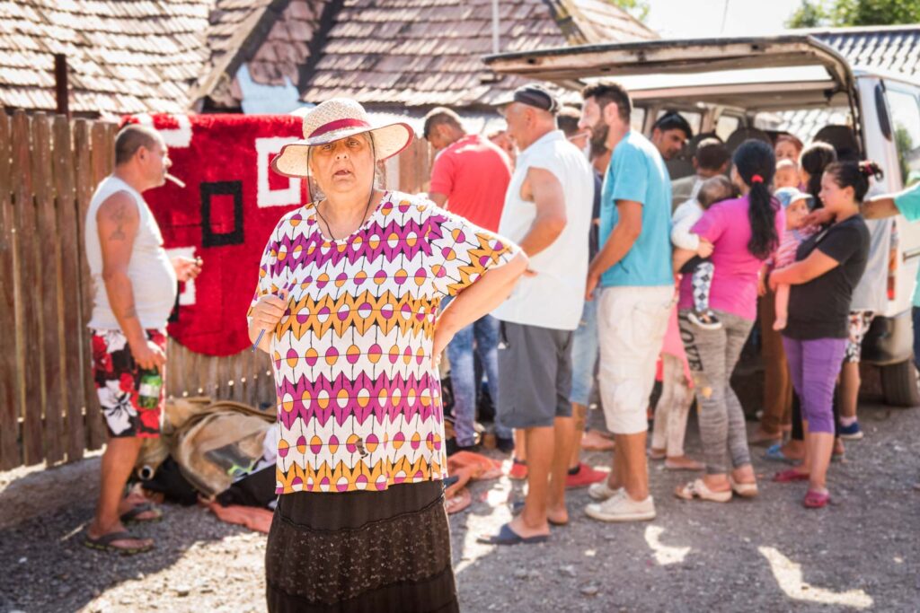 Eine Frau mit buntem Oberteil und Strohhut steht im Vordergrund, während im Hintergrund eine Gruppe von Menschen bei einem Fahrzeug versammelt ist.