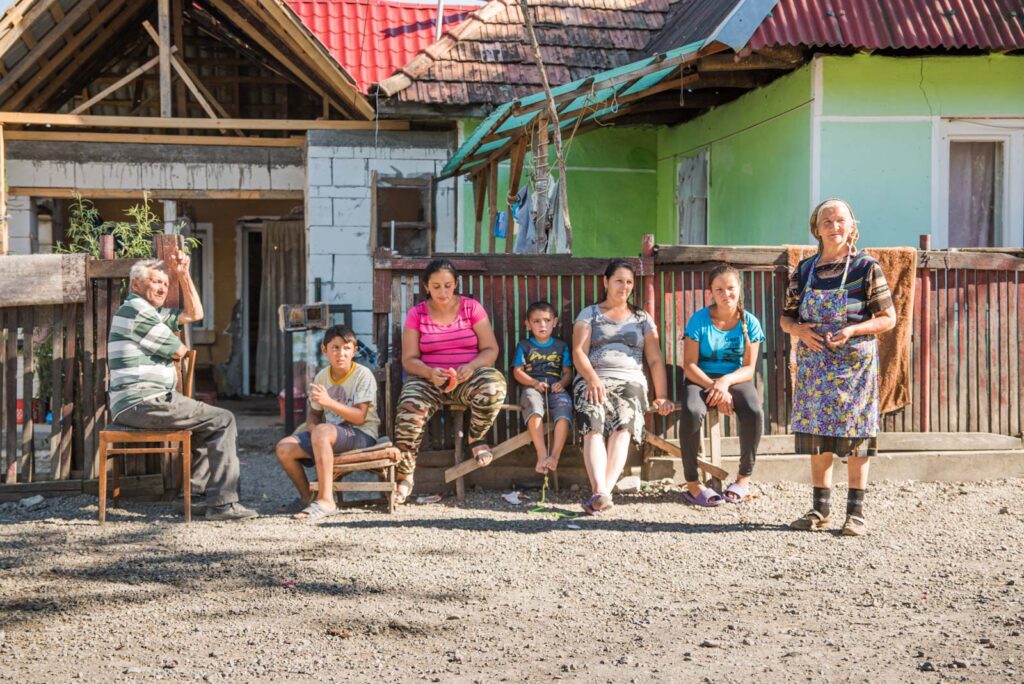 Mehrere Personen sitzen vor einem Haus auf Stühlen und Holzbänken, eine ältere Frau steht daneben. Das Haus ist teilweise mit Ziegeln bedeckt und hat eine grüne Wand, die Gruppe wirkt entspannt und in ein Gespräch vertieft. Der Boden ist aus Kies.