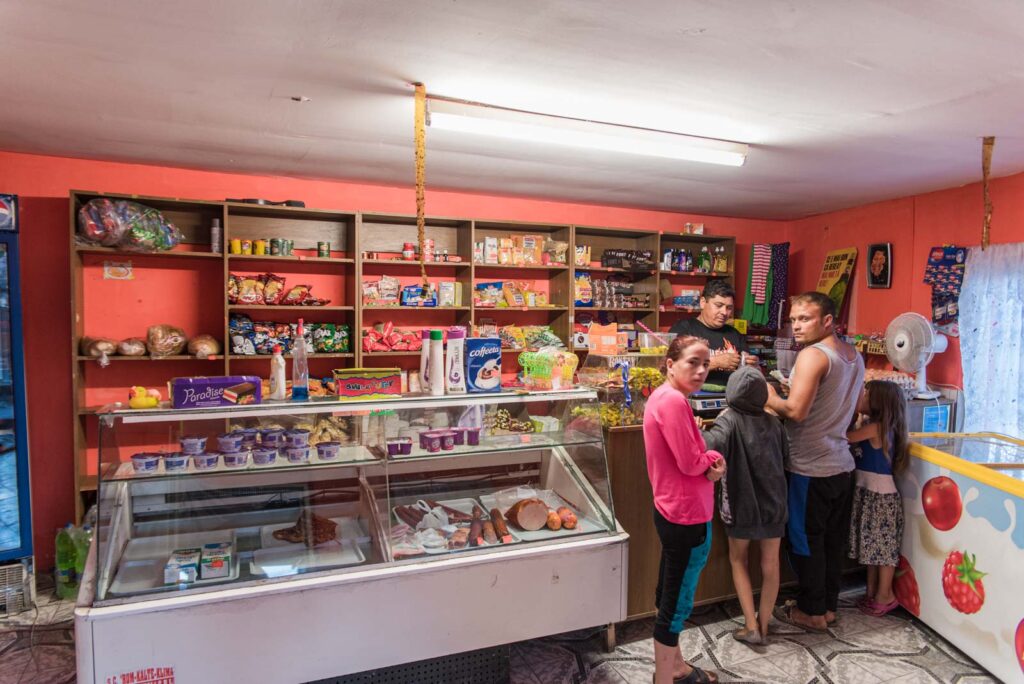 Ein kleiner Laden mit einer Theke voller Snacks, Getränken und verschiedenen Produkten in Regalen. Mehrere Personen stehen an der Kasse, darunter eine Frau im rosa Pullover, ein Mann im ärmellosen Shirt und Kinder. An der Wand befinden sich dekorative Gegenstände und Farbakzente.