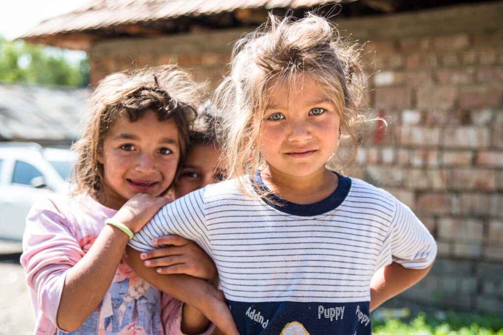 Drei Kinder stehen draußen vor einem Gebäude, zwei Mädchen im Vordergrund und ein weiteres Kind lächelt im Hintergrund zwischen ihnen hindurch.