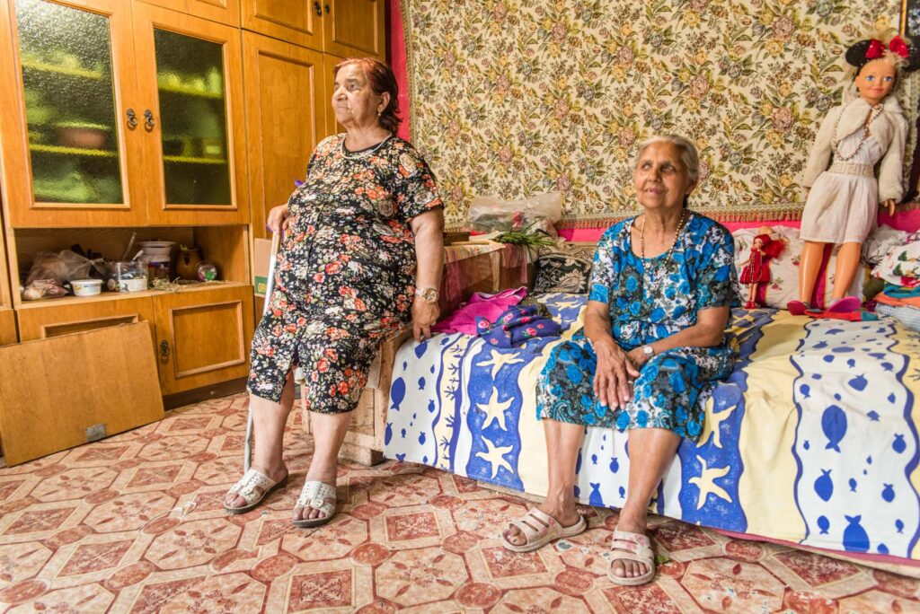 Zwei ältere Frauen sitzen in einem farbenfrohen, gemütlichen Raum mit gemusterten Tapeten und einem Holzschrank. Eine Frau sitzt auf einem Bett mit buntem Bettüberwurf, die andere lehnt an einem Karton. Im Hintergrund sind Puppen zu sehen.