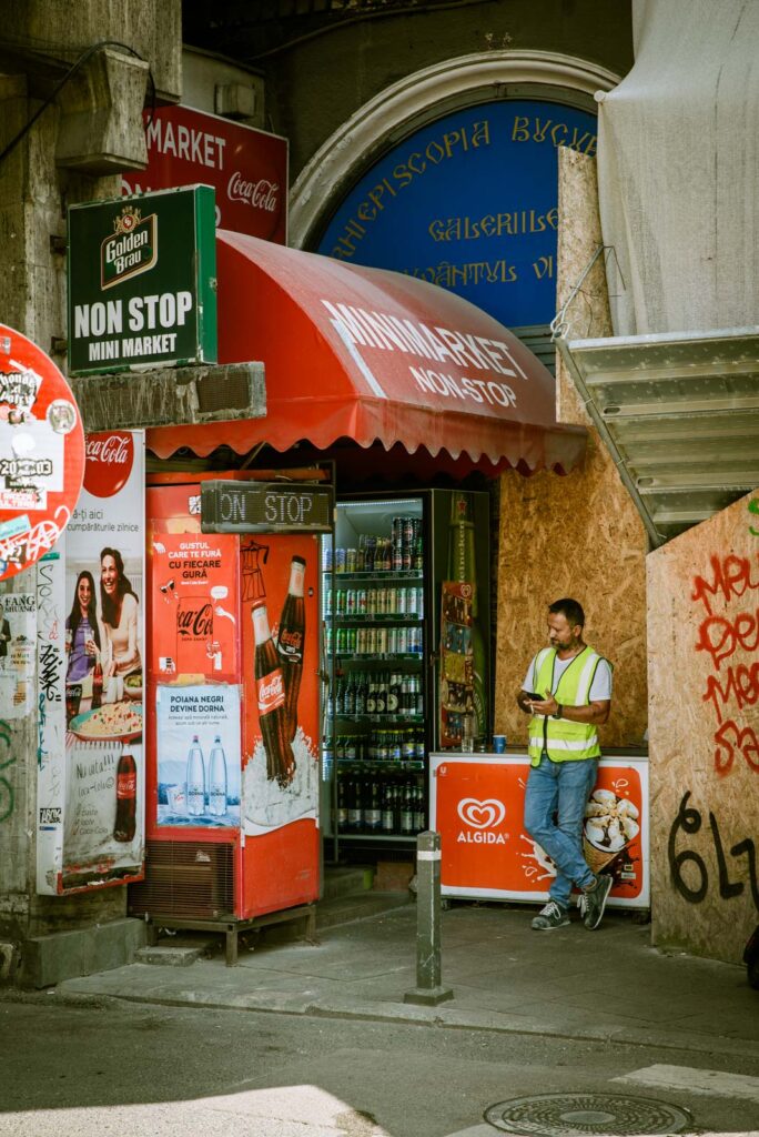 Ein Mann in gelber Warnweste steht vor einem Mini-Market mit rotem Vordach, umgeben von Werbeplakaten für Coca-Cola und Algida-Eiscreme, während er auf sein Mobiltelefon schaut.