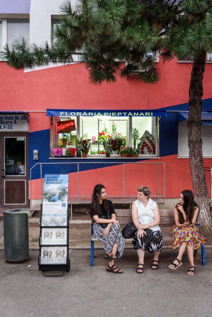 Drei Frauen sitzen auf einer Bank vor einem Blumenladen mit der Aufschrift 'FLORĂRIA PIEPTANARI'. Der Laden hat eine auffällige Fassade in Rot und Blau und im Schaufenster sind verschiedene Blumen ausgestellt. Auf dem Boden steht ein Schaukasten mit religiösem Material.
