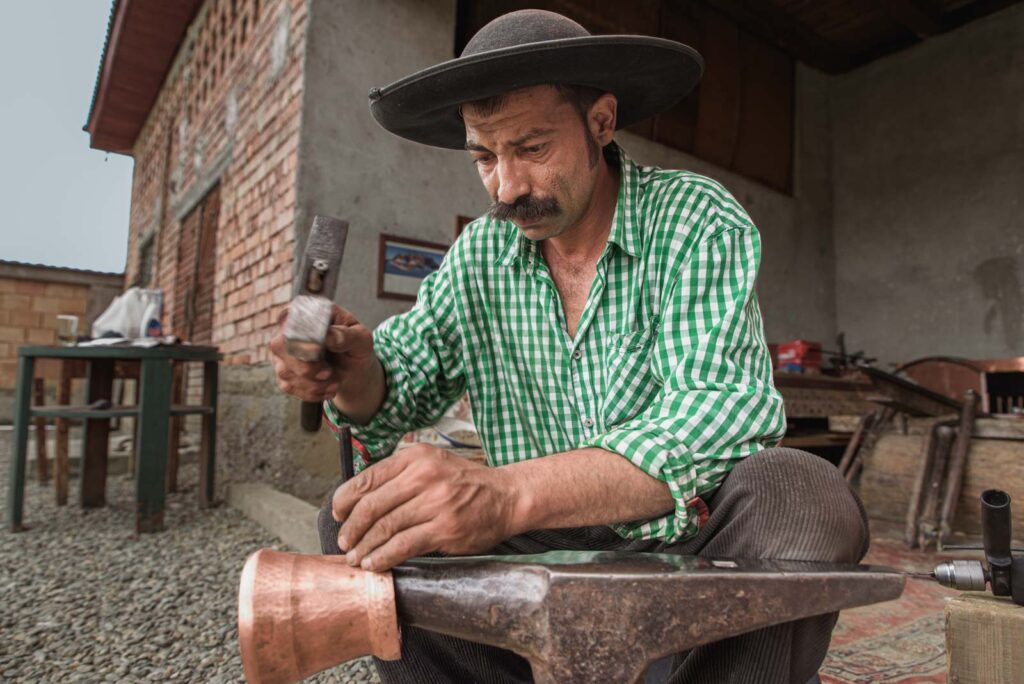 Ein Mann mit einem breitkrempigen Hut und einem karierten Hemd bearbeitet ein Metallstück mit einem Hammer auf einem Amboss.