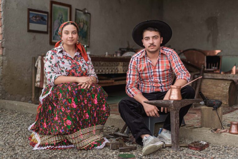 Ein Mann und eine Frau in traditioneller Kleidung sitzen in einer Werkstatt, umgeben von Werkzeugen und einem Amboss mit einem Kupferkrug darauf.